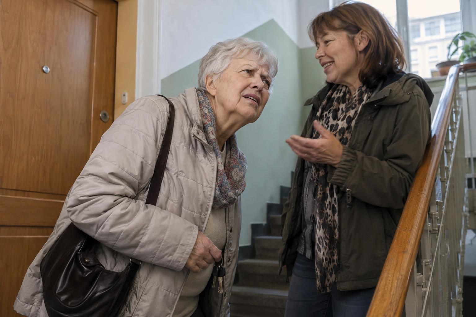 Starejša ženska z naglušnostjo se na stopnišču bloka s ključi v roki nagne k sosedi, ki ji nekaj pripoveduje, in negotovo dvigne obrvi